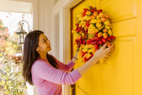 A woman putting a red and yellow tulip flower wreath on a yellow front door.
