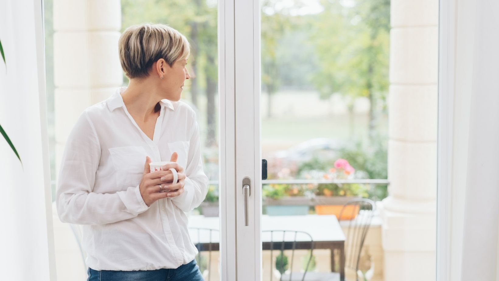 A woman holding a mug and looking out a patio door.